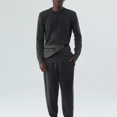 Homem com conjunto escuro casual, cinto saco verde-oliva e calçado preto diante de fundo branco.