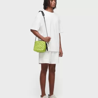 Homem com roupa branca e ténis com bolsa verde em fundo cinza claro