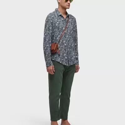 Homem com camisa azul escura, calças verdes, sandálias pretas, óculos amarelos e bolsa castanha