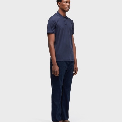 Homem com polo azul escuro, calças azul escuro e sandálias castanhas em fundo cinzento.