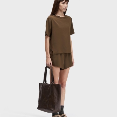 Mulher com conjunto castanho, calções, t-shirt, mala de pele castanha, meias e sandálias castanhas.