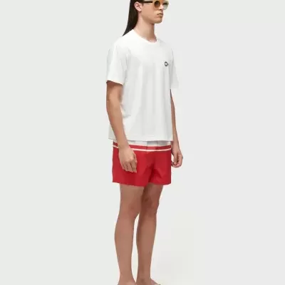 Homem com camisola branca, calção vermelho com riscas brancas, chinelos brancos e óculos de sol amarelos.