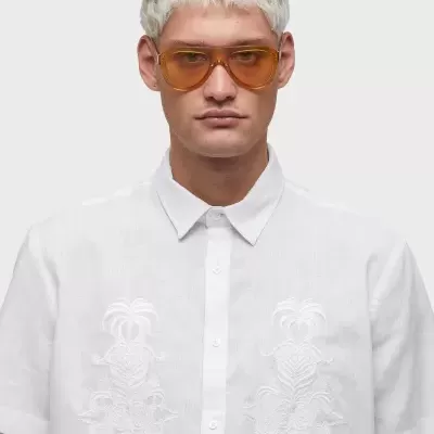 Homem de cabelo platinado a usar camisa branca com bordados e óculos de sol castanhos com lentes laranja.