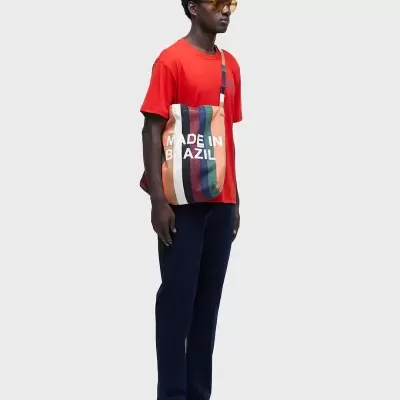 Homem com t-shirt vermelha, calças azuis escuras, sandálias negras, óculos de sol amarelos e saco de tecido às riscas coloridas