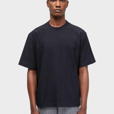 Homem de pele escura com t-shirt preta e calças cinzentas em fundo branco.