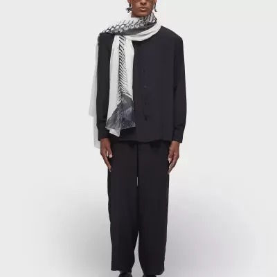 Homem vestido com camisa preta, calças pretas largas, sapatos pretos, écharpe preta e branca e óculos escuros