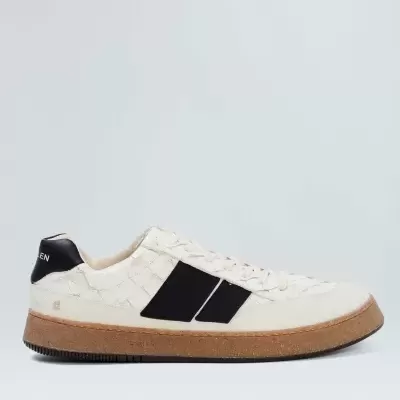 Tênis branco com detalhes pretos e sola castanha em fundo branco.