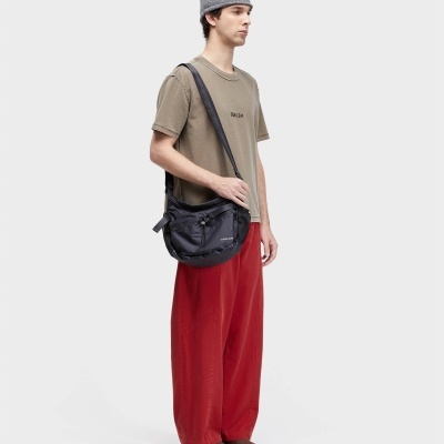 Homem veste t-shirt bege OSKLEN, calças vermelhas largas, gorro cinza e mala preta OSKLEN ao ombro.