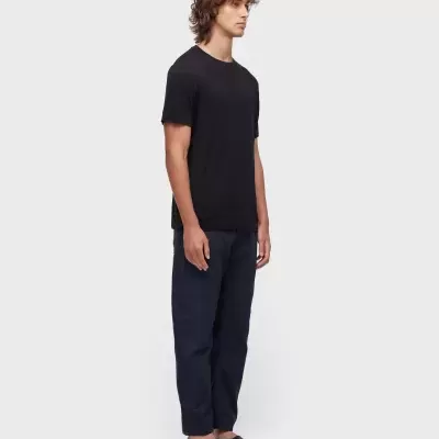 Homem com t-shirt preta, calças escuras e sandálias pretas, em fundo cinzento claro