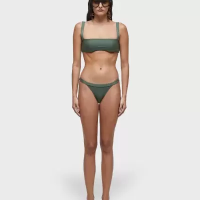 Mulher com biquíni verde musgo, sandálias pretas e óculos escuros em fundo branco