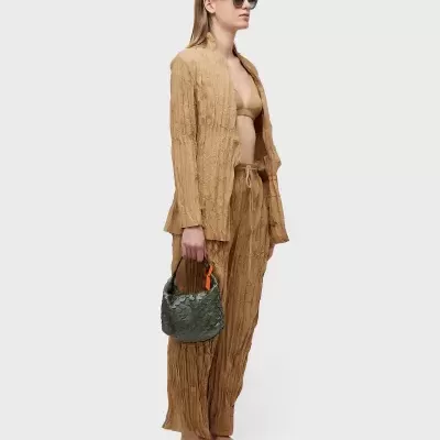 Mulher vestindo blazer e calças castanhas texturizadas, top castanho, sapatos brancos, segurando mala cinzenta com pega laranja em fundo cinzento claro.