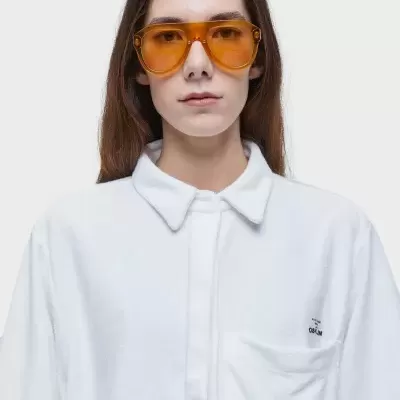 Pessoa com óculos de sol de armação larga e lentes laranja, vestindo camisa branca com bolso e gola clássica.