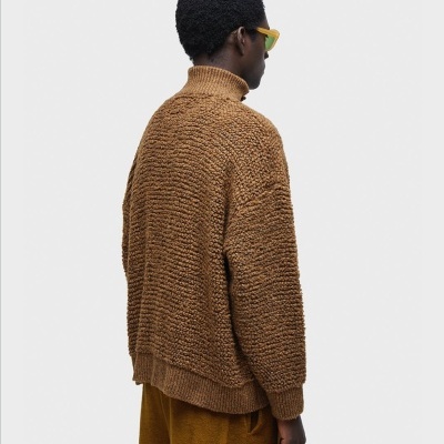 Homem de costas com camisola castanha de gola alta e calças largas castanhas