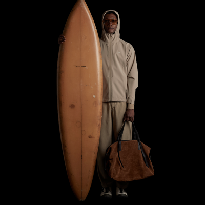 Homem com casaco bege e prancha de surf castanha, segura bolsa em camurça castanha