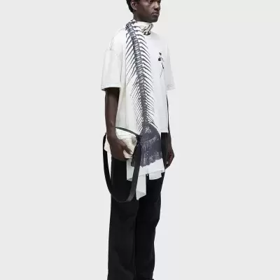 Homem vestindo t-shirt branca estampada, calças pretas e segurando mala bege.