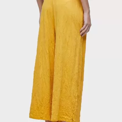 Calças largas amarelas texturizadas com padrão floral, usadas por trás, com sandálias douradas em fundo cinzento claro