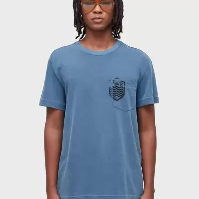 Camiseta azul com bolso e emblema preto no peito e calções azuis usada por modelo com óculos de sol