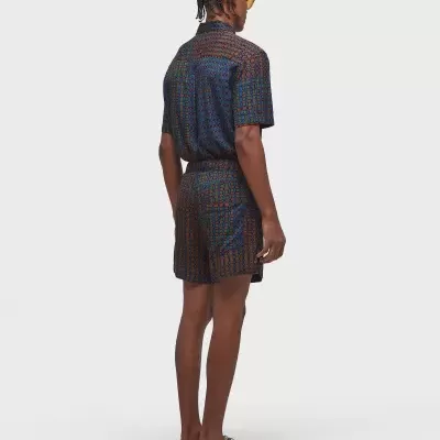 Homem com camisa e calções estampados coloridos, sapatos bege e óculos amarelos, visto de costas