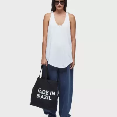 Mulher com regata branca, calças azuis, ténis brancos, óculos escuros e mala preta com texto MADE IN BRAZIL