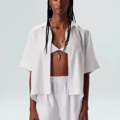 Modelo feminina com conjunto branco composto por camisa, top e calções, em fundo neutro.