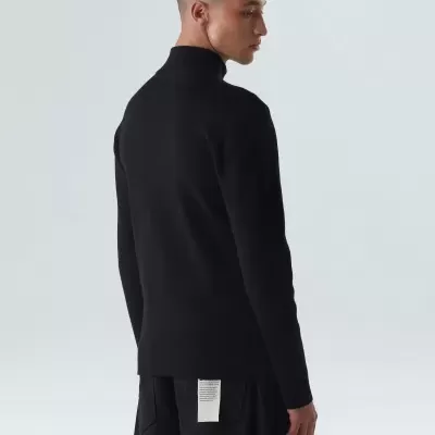 Homem de costas com camisola preta de gola alta e calças pretas largas em fundo neutro.