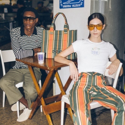 Duas pessoas sentadas com roupa e mala às riscas verdes, laranja e bege junto a mesa de madeira