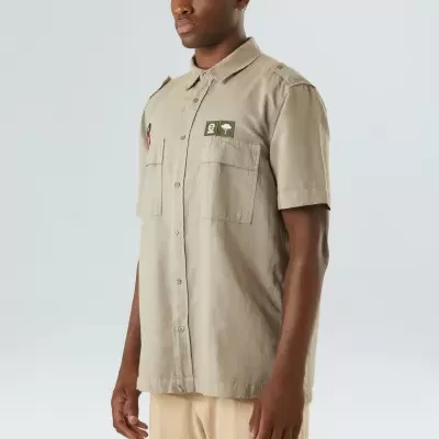 Camisa bege de manga curta com dois emblemas verdes no peito, vestida por modelo masculino