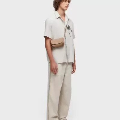 Homem com roupa bege casual com estampa de palmeira e bolsa cruzada.