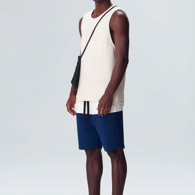 Homem com camisola branca de alças, calções azuis, chinelos pretos e bolsa preta em fundo branco.