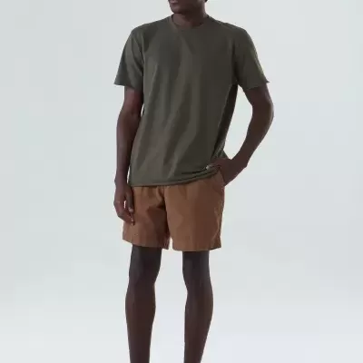 Homem com t-shirt verde-oliva, calções castanhos e ténis verdes em fundo cinzento claro