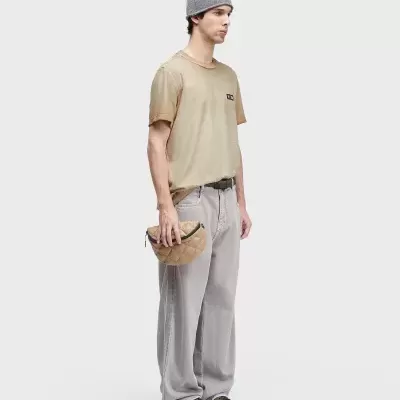 Homem com t-shirt bege, calças cinza largas, boné cinza, bolsa bege acolchoada e sapatilhas castanhas com branco