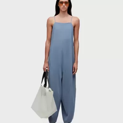 Mulher com macacão azul claro e óculos de sol, segurando saco branco com alças pretas