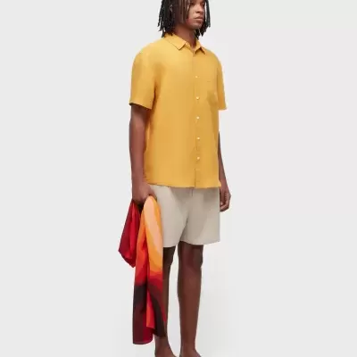 Homem com camisa amarelo mostarda, calções bege e sandálias pretas segurando tecido colorido