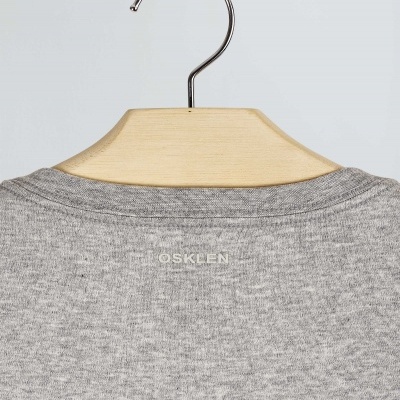 Peça de vestuário cinza claro pendurada num cabide de madeira com texto OSKLEN