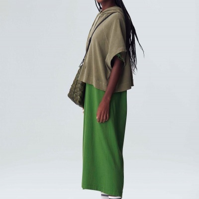 Mulher com vestido verde comprido, capa verde-oliva, sapatilhas brancas com texto, meias brancas, bolsa verde escura, fundo branco.