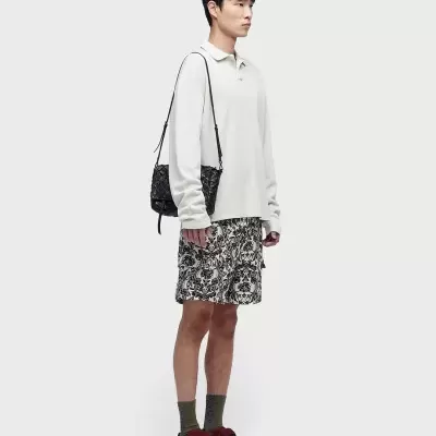 Homem com roupa casual: camisola branca, calções florais, meias cinzentas e sapatos bordô, com mala preta ao ombro