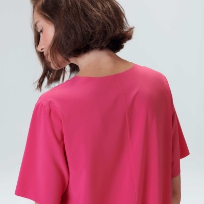 Top rosa vibrante de mangas curtas visto de trás com modelo de cabelo castanho curto, fundo cinza.