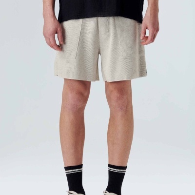 Shorts bege claros masculinos com textura, meias pretas com riscas brancas e ténis brancos em fundo neutro.