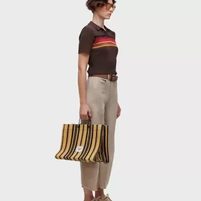 Modelo com camisa castanha de malha, calças claras, ténis bege e mala listrada amarela e preta