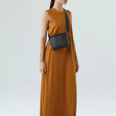 Mulher a usar vestido comprido castanho alaranjado, ténis brancos e bolsa preta pequena de tecido com alça larga, fundo cinzento