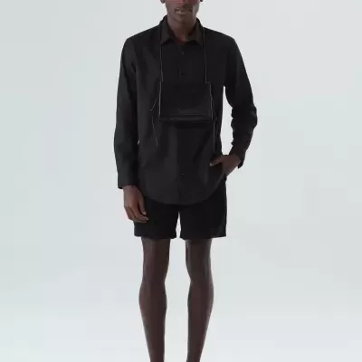 Modelo masculino com camisa preta, calções pretos, sapatos pretos e bolsa preta pendurada no pescoço