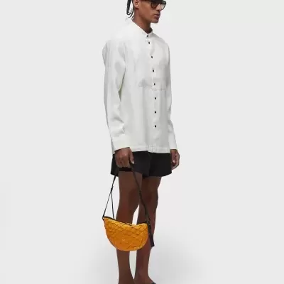 Homem com camisa branca, calções pretos, sandálias, óculos escuros e bolsa amarela trançada.