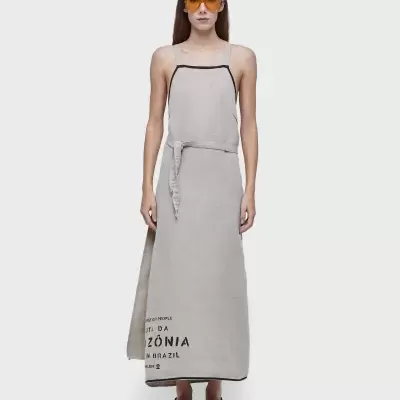 Mulher com vestido comprido cinzento claro com alças finas e texto preto, óculos de sol laranja e sandálias pretas