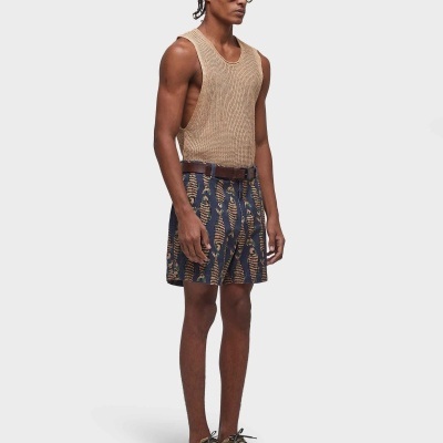 Homem com colete bege, calções azuis com padrão castanho, cinto, ténis bege e óculos escuros