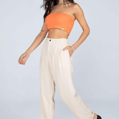 Mulher com top laranja e calças bege claro com sandálias pretas em fundo cinza