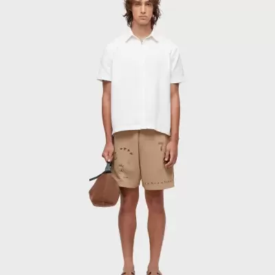 Homem com camisa branca, calções bege decorados, bolsa castanha e sandálias bege