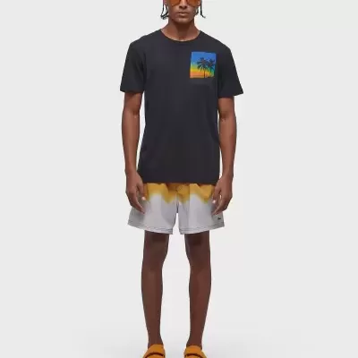 Homem com t-shirt preta com estampa de palmeiras, calções brancos com degradê amarelo e laranja, óculos de sol castanhos e sandálias laranjas