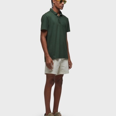 Homem com polo verde e calções brancos, calçado verde e óculos de sol amarelos