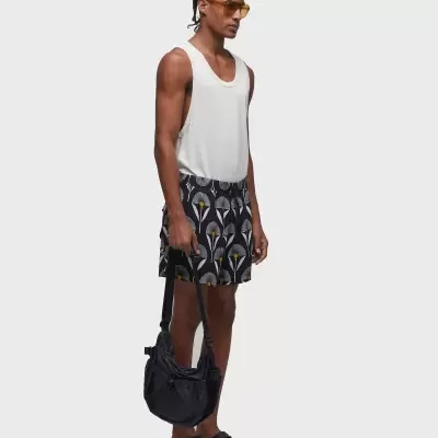 Homem com camisola branca, calções pretos com padrão floral, sandálias pretas, óculos amarelos e mala preta