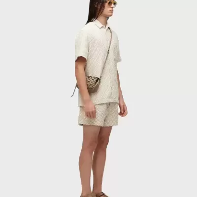 Homem com roupa bege texturizada, sandálias castanhas, óculos amarelos e bolsa castanha pequena.